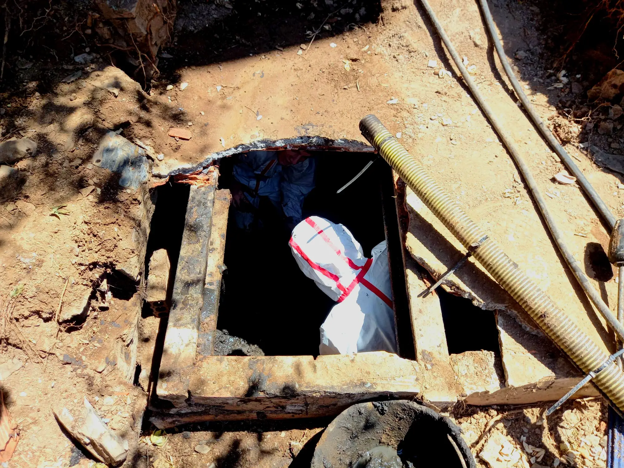 Operario con traje de protección blanco trabajando dentro de un pozo de registro del alcantarillado municipal, con la manguera de aspiración del camión cuba al lado